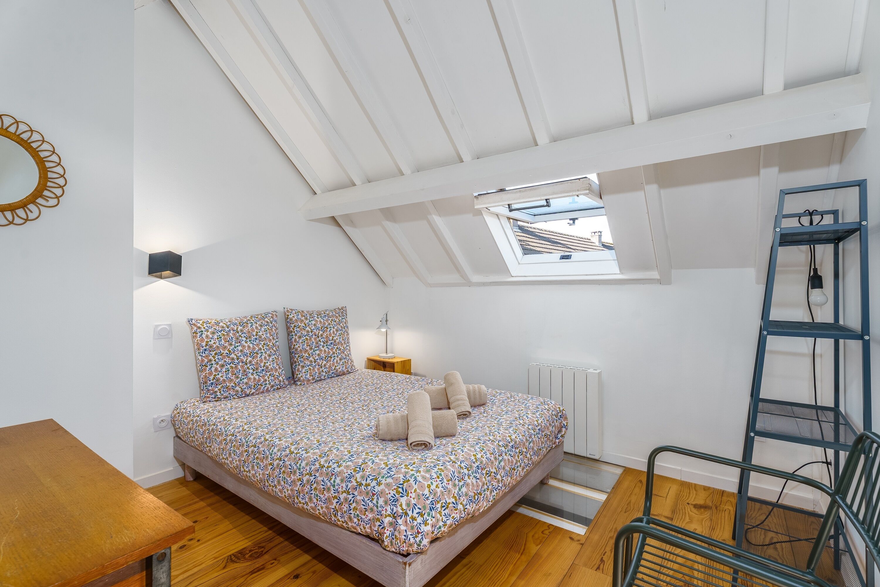 Une chambre avec un plafond en pente, un puits de lumière, un lit avec une literie à motifs, une table de chevet, une lampe, un radiateur, un plancher en bois et une chaise en métal.