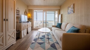 A living room with a sofa, a coffee table, a TV, a desk with a lamp, and a view through a glass door. - Les Essarts Marine - Au bord de la plage (Courseulles-sur-Mer)