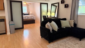 Una sala moderna con un sofá negro, un tapete pequeño, piso de madera, una cama con ropa de cama estampada, un escritorio con una lámpara y un espejo grande.