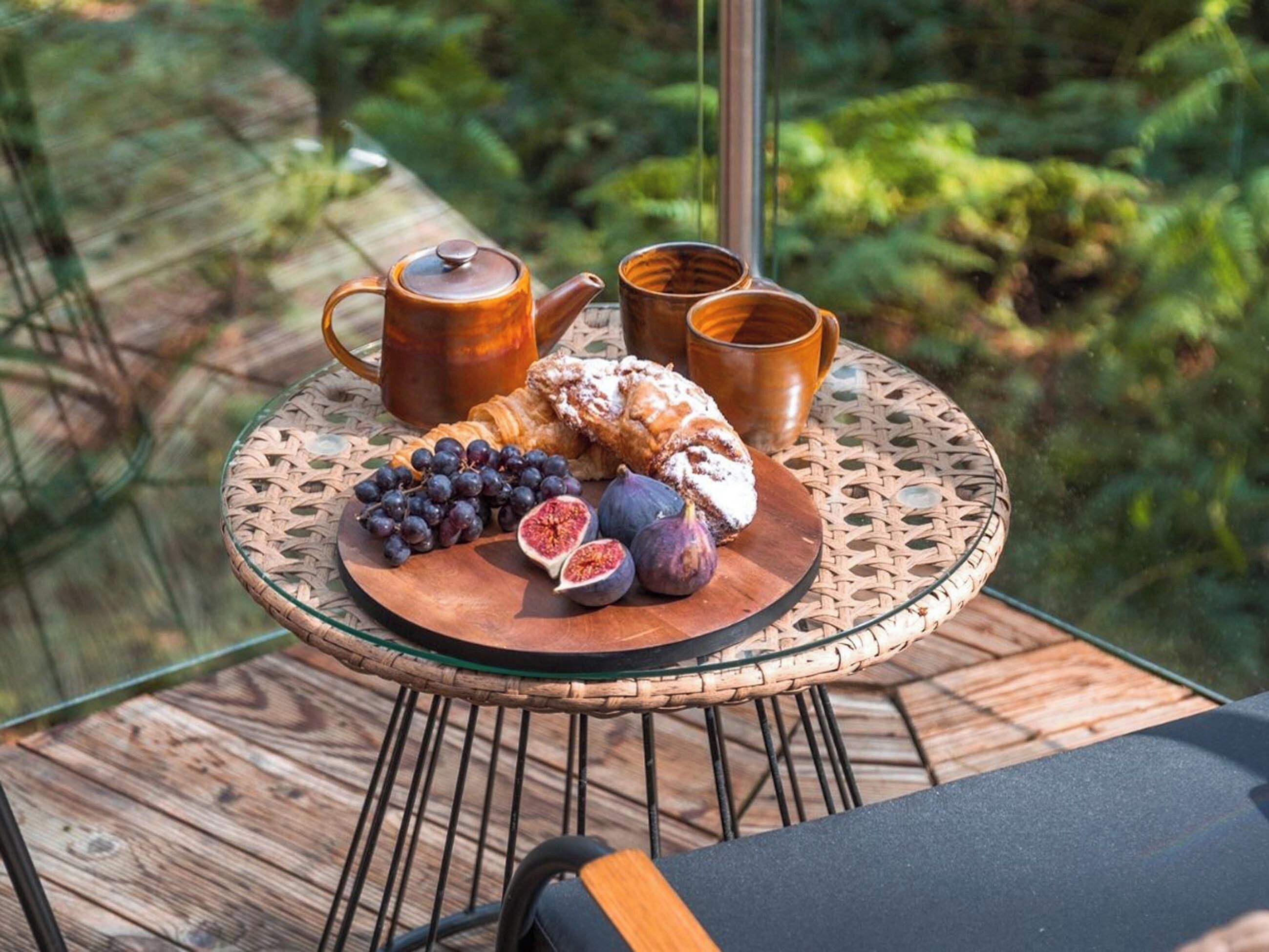 A round glass table with a woven edge, a teapot, two cups, a plate with grapes, figs, and pastries on a balcony with a wooden railing and greenery in the background.