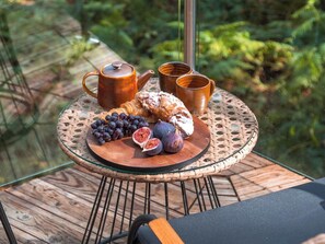 Una mesa redonda de vidrio con borde tejido, una tetera, dos tazas, un plato con uvas, higos y pasteles en un balcón con barandilla de madera y follaje al fondo.