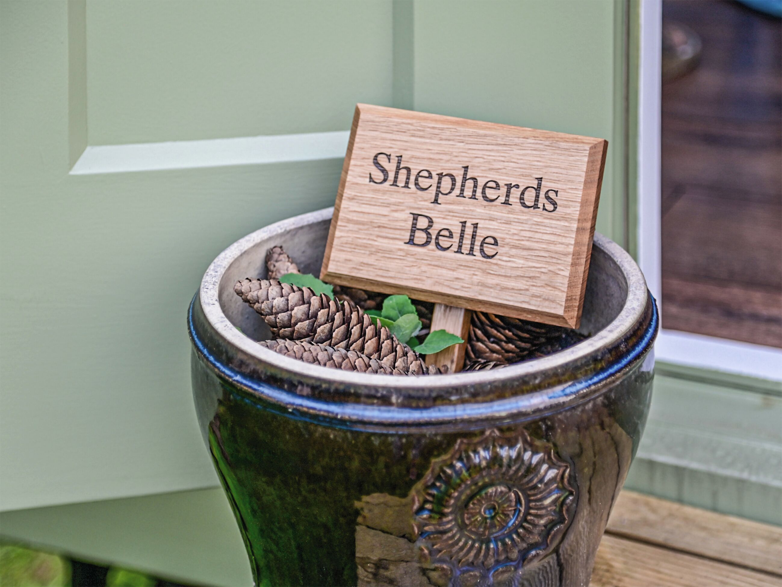 A ceramic pot with a wooden sign reading 'Shepherds Belle' and pinecones inside.