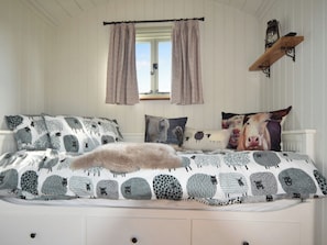 Une chambre à coucher avec un lit, des oreillers, une fenêtre avec des rideaux, une étagère fixée au mur avec une lanterne, et une porte blanche.