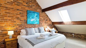 A bedroom with a brick wall, a bed with white bedding, a blue painting, and a skylight. - Spacious stone barn close to Matlock and Carsington Water. (Carsington, near Ashbourne)