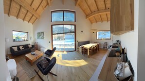 A spacious living area with a high ceiling, wooden beams, a sofa, a coffee table, a dining table with chairs, and large windows offering a view of the outdoors. - Le Chalet Morets - Avec vue sur la montagne (Saint-Gervais-les-Bains)
