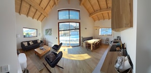 Un vaste espace de vie doté d’un haut plafond, de poutres en bois, d’un canapé, d’une table basse, d’une table à manger avec des chaises, et de grandes fenêtres offrant une vue sur l’extérieur.