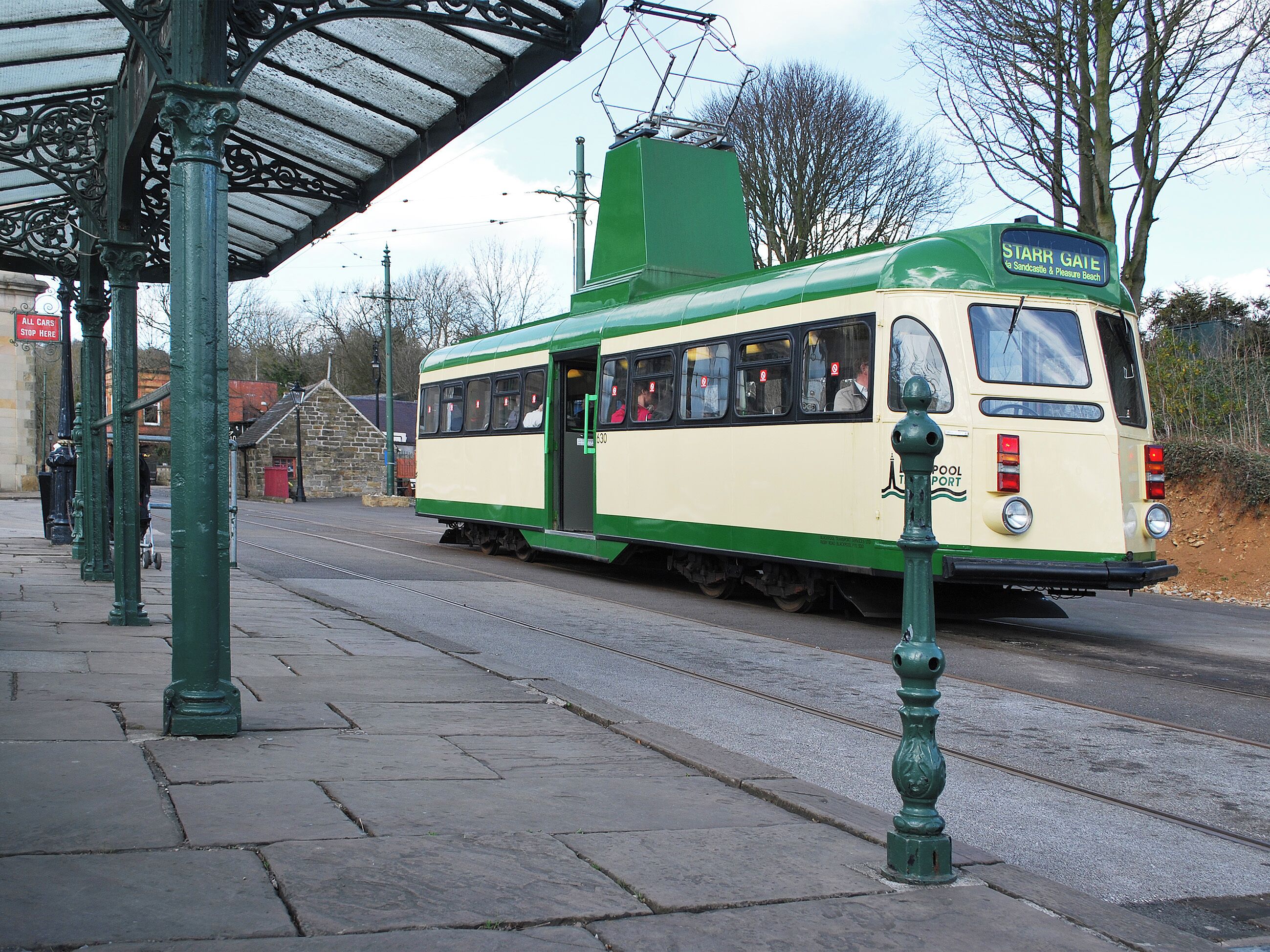 A green and white tram at a station with a covered waiting area and stone pavement.