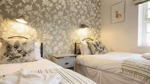 A bedroom with two beds, floral wallpaper, bedside lamps, a nightstand, a framed picture, and a window. - Spacious stone cottage set in beautiful woodland with lovely views over a lake. (Darwin Lake, near Matlock)