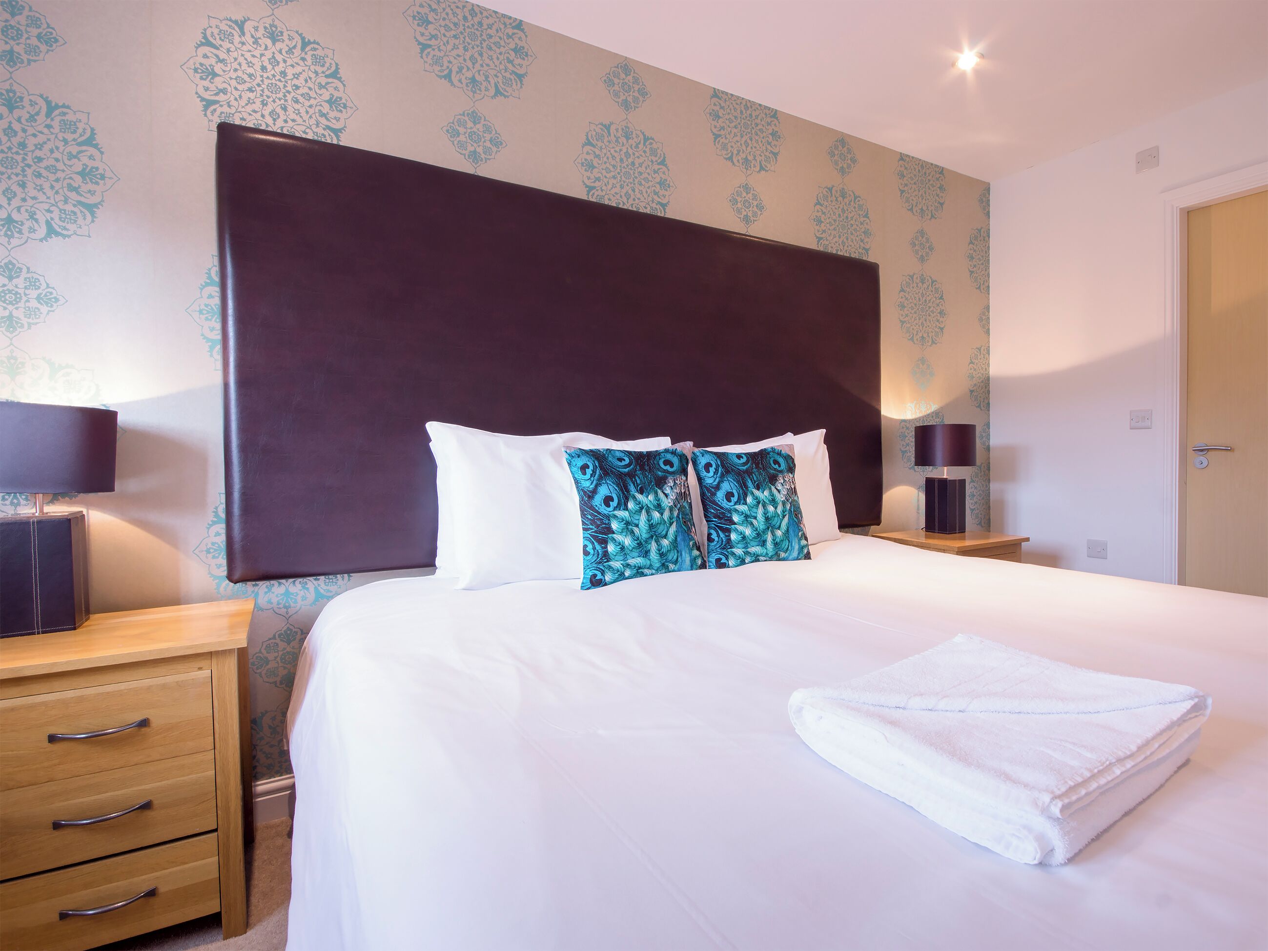 A neatly made bed with white and blue pillows, a folded white towel, and a dark headboard. There are two bedside tables with lamps, a wooden nightstand, and a door to the right.