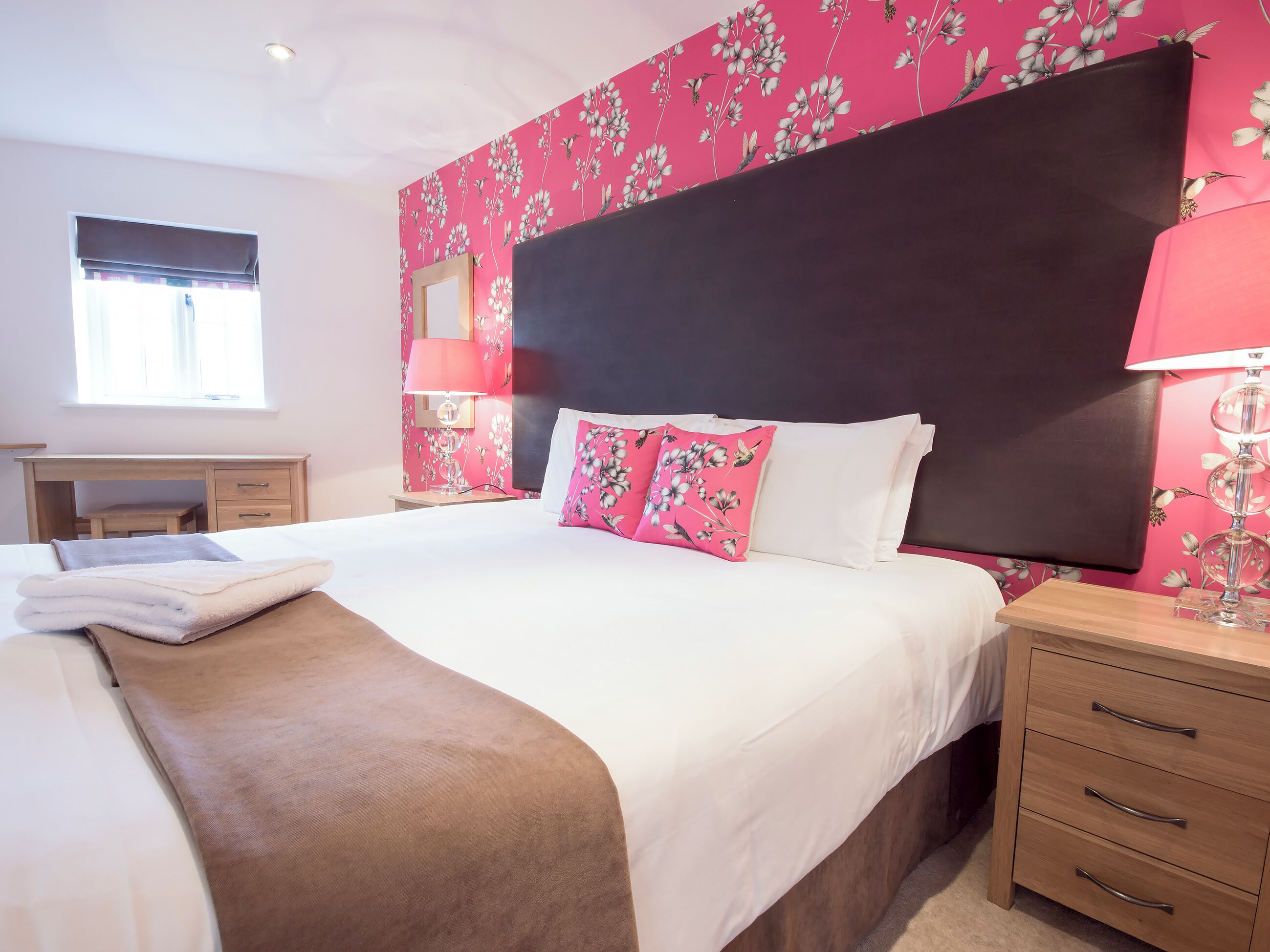 A hotel room with a bed, bedside tables, a desk, and a window.