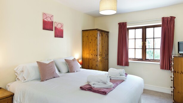 A neatly made bed with pillows, a wooden wardrobe, a window with curtains, and a hanging light fixture. - Attractive barn conversion set in three acres of shared grounds. (Carsington, near Ashbourne)