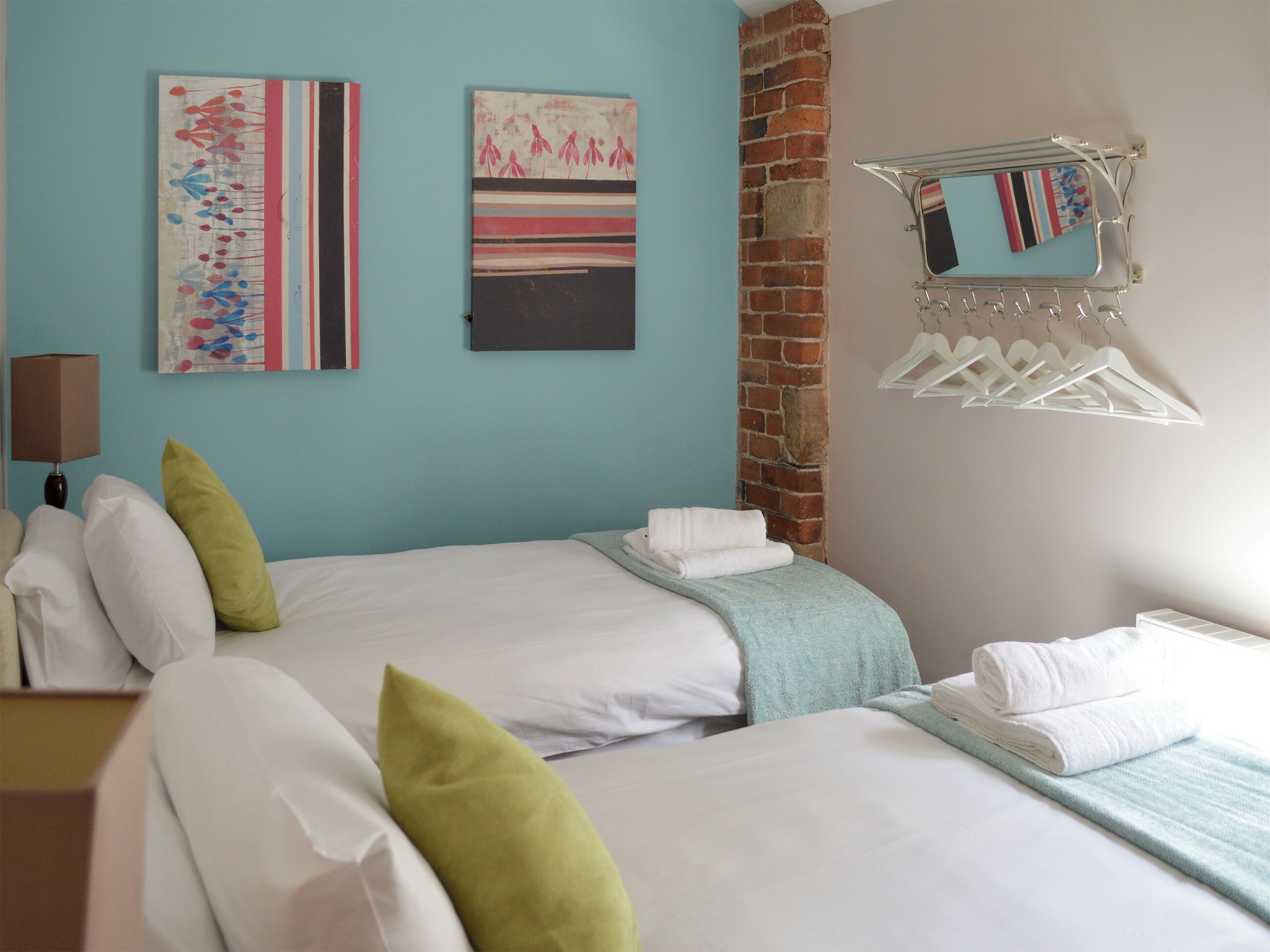 A hotel room with two beds, a brick wall, a wall-mounted towel rack, and abstract art on the wall.