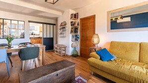 A living room with a sofa, a dining table, and a kitchen in the background. - L'Orangerie - Terrasse avec vue sur la montagne (Ajaccio)