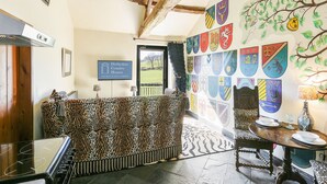 A room with a wooden ceiling, a sofa with a leopard print cover, a dining table with a vase, and a wall decorated with various coats of arms.