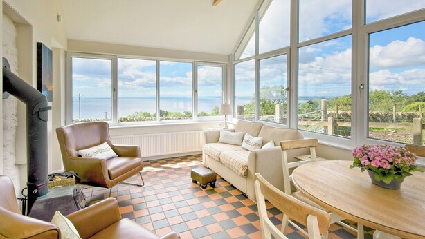 A sunlit room with a large window, a sofa, a chair, a table, and a view of the sea.