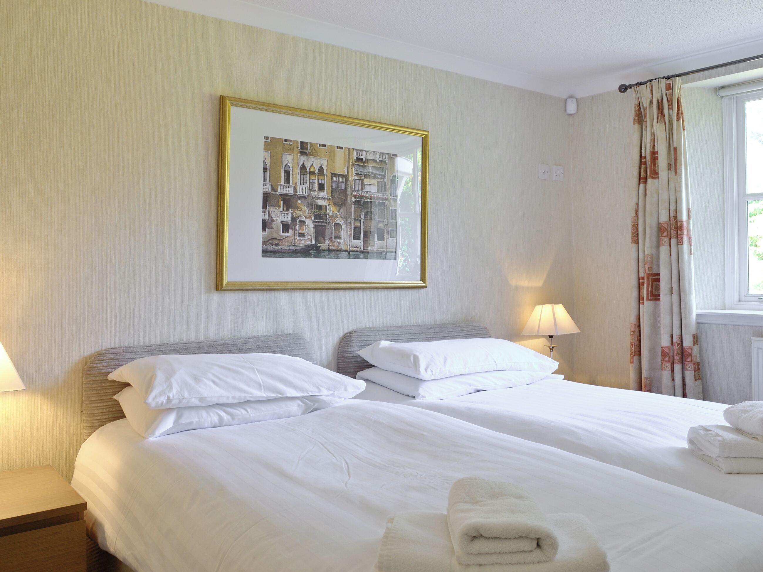 A hotel room with two beds, a framed picture on the wall, a window with curtains, and a bedside lamp.