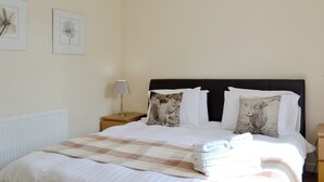 Een net opgemaakt bed met een geruit plaid, een bedlampje en twee ingelijste botanische prints aan de muur.