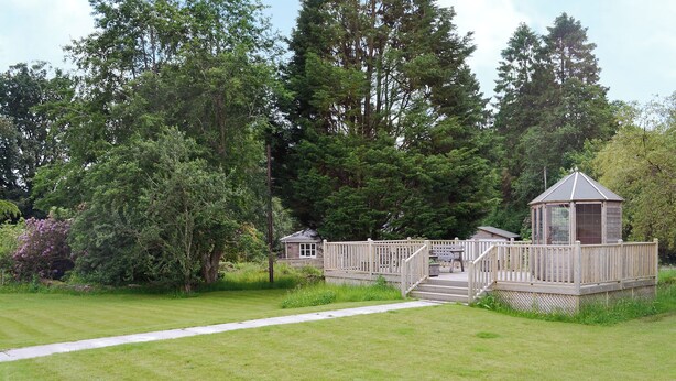 Een houten terras dat leidt naar een pergola met een klein gebouw, omgeven door weelderige begroeiing en bomen.