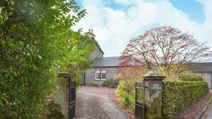 Une charmante maison en pierre avec une allée de gravier menant à une entrée fermée par un portail, le tout entouré d’arbres et d’arbustes.