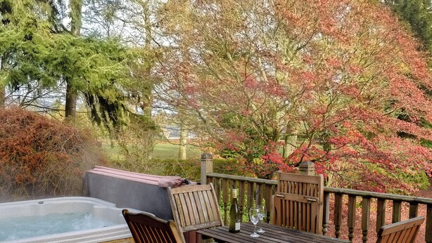 Un bain à remous sur une terrasse en bois, agrémentée de chaises et d’une table, le tout entouré d’arbres aux feuilles d’automne.