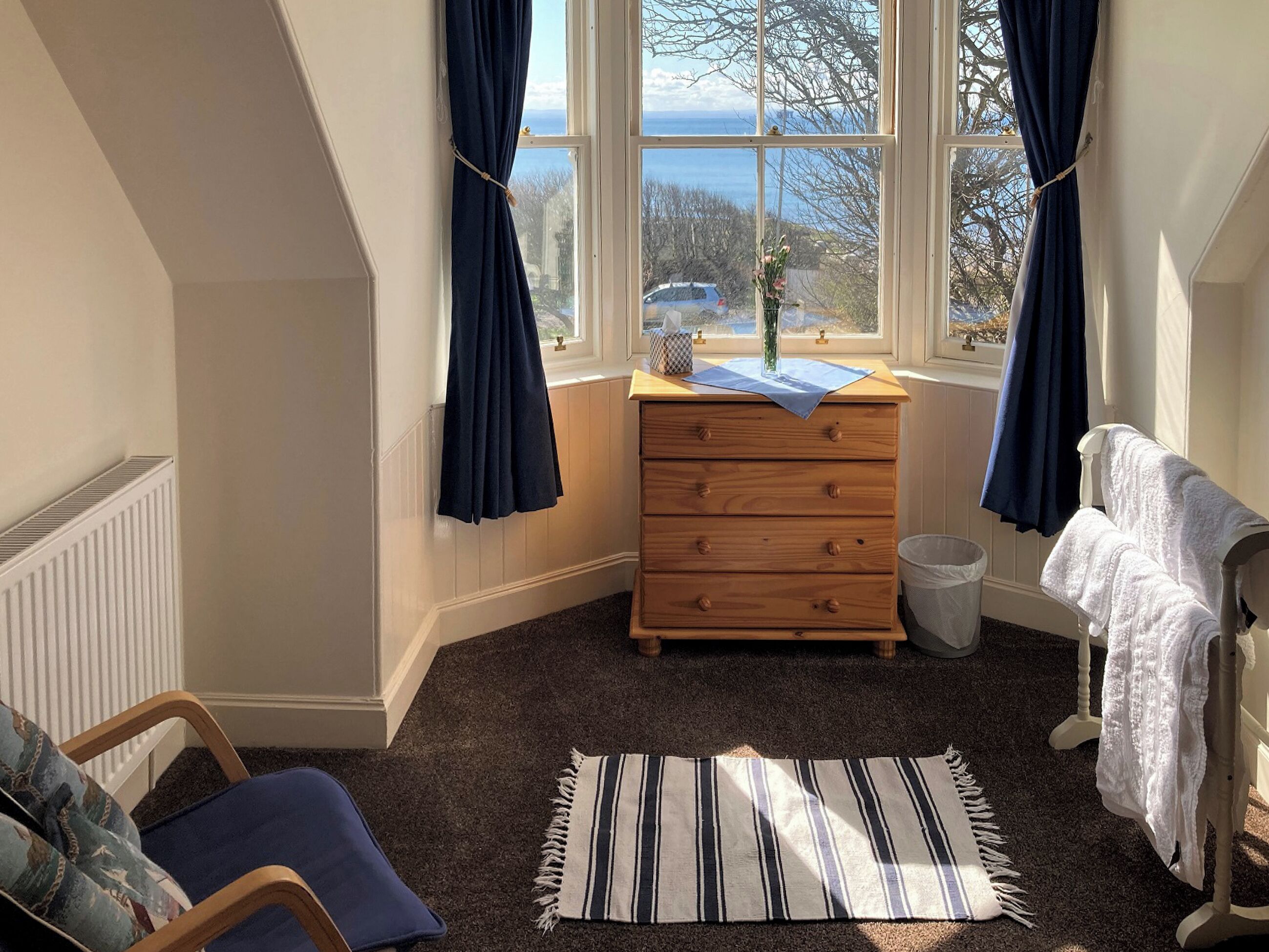 A bedroom with a window, a wooden dresser, a chair, a radiator, a rug, and a towel rack.