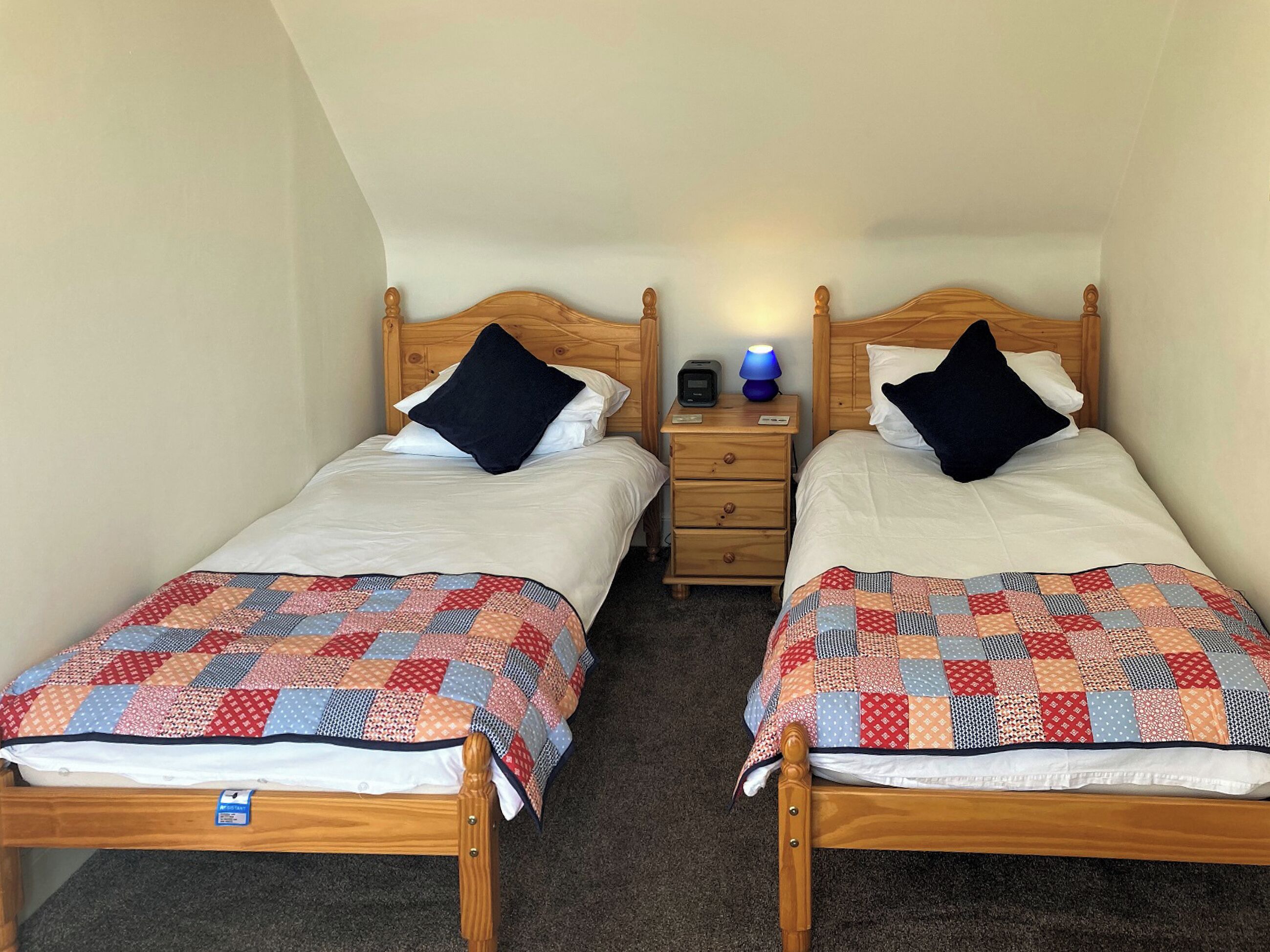 A twin room with two wooden beds, each with a quilt and pillow, a nightstand with a lamp, and a small table between the beds.