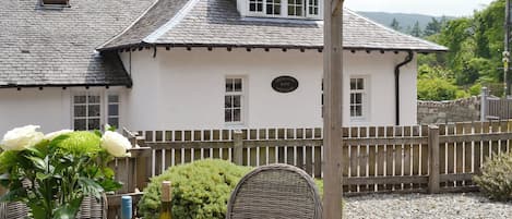 Une petite maison blanche avec une terrasse en bois, une table en osier ornée de bouteilles de vin et un chemin de gravier.