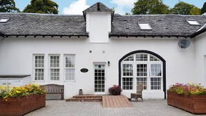 Een witte cottage met een geruit trottoir ervoor, een bankje en bloembakken.