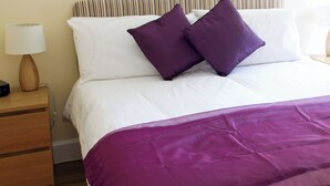 Una cama con ropa blanca y dos almohadas moradas, una mesita de noche de madera con una lámpara, y un plaid morado.