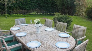 Une table ronde en bois, dressée pour six personnes, avec des chaises assorties, le tout entouré d’un jardin verdoyant.