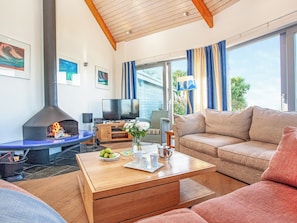 Ein Wohnzimmer mit Kamin, Fernseher, einer Couch, einem Couchtisch und Blick ins Freie.