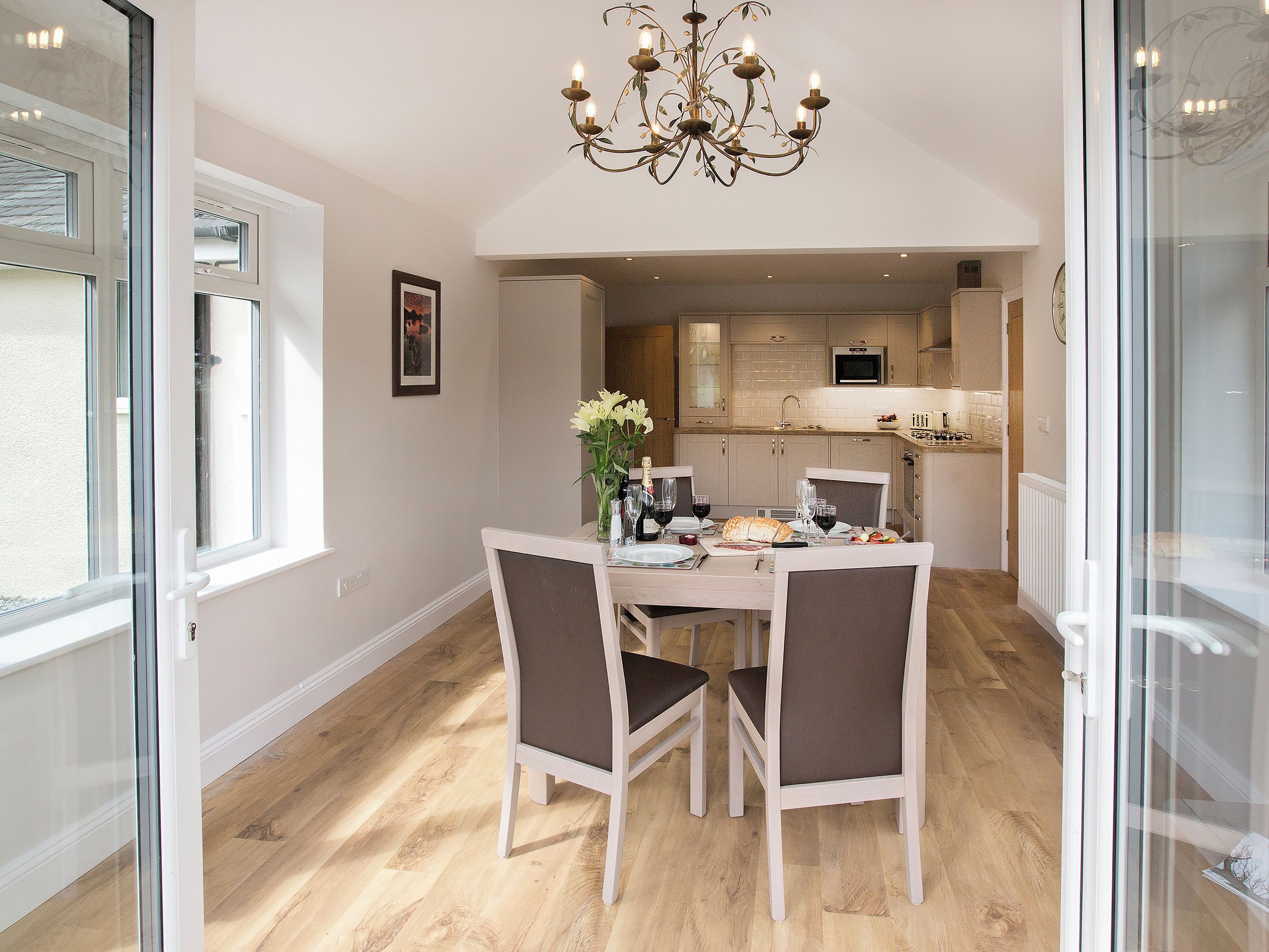 A dining area with a chandelier, a table set for four, and a kitchen in the background.