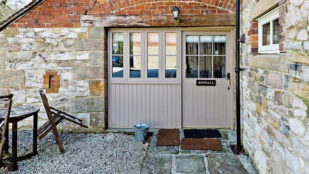 A stone building with a wooden door labeled 'Bonsall,' a small window, and a metal bucket on the ground. - Bonsall Cottage - RDDD (Carsington, near Ashbourne)