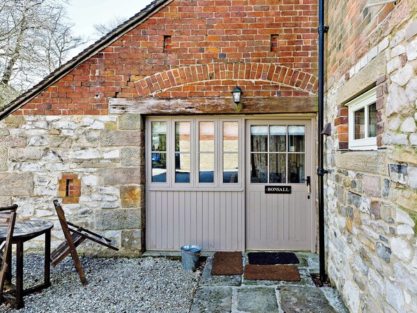 A stone building with a wooden door labeled 'Bonsall,' a small window, and a metal bucket on the ground. - Bonsall Cottage - RDDD (Carsington, near Ashbourne)