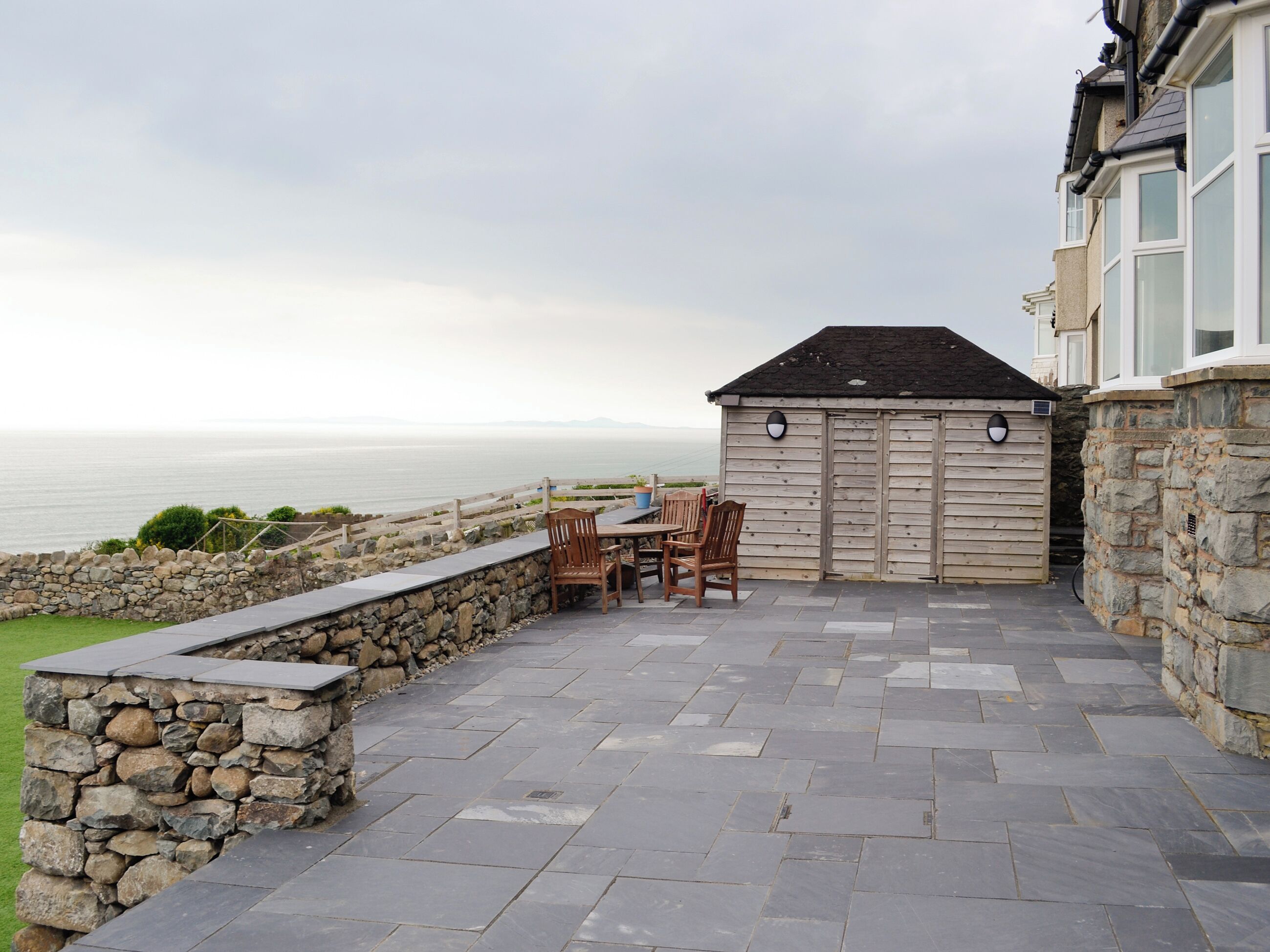 A patio with stone walls, a wooden table, and chairs overlooking the sea.