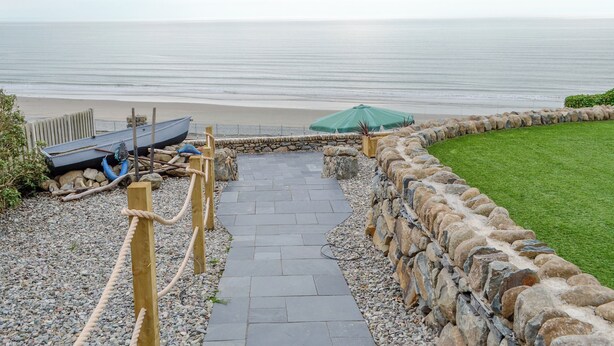 Un sentiero in pietra conduce a una spiaggia, con una recinzione in legno, un ombrellone verde e una barca.