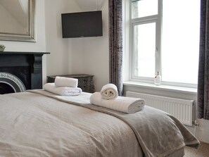 A neatly made bed with rolled towels, a flat-screen TV mounted on the wall, a fireplace, and a window with curtains.