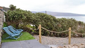 Un muro in pietra, una zona erbosa con poltrone da relax blu, una barriera in corda e vista sul mare.