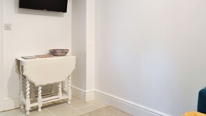 A white room with a television mounted on the wall, a small white table with a colorful bowl and magazines, and a blue chair in the corner. - Brig-Y-Don Bach - UK11309 (Llanaber, near Barmouth)