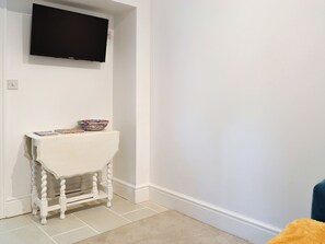 A white room with a television mounted on the wall, a small white table with a colorful bowl and magazines, and a blue chair in the corner. - Brig-Y-Don Bach - UK11309 (Llanaber, near Barmouth)