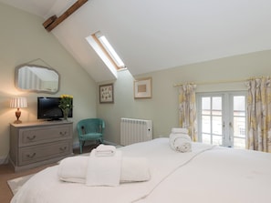 A bedroom with a bed, a dresser, a television, a chair, a radiator, and a window with curtains. - No.2 Meynell Mews - UK11365 (Breedon on the Hill)