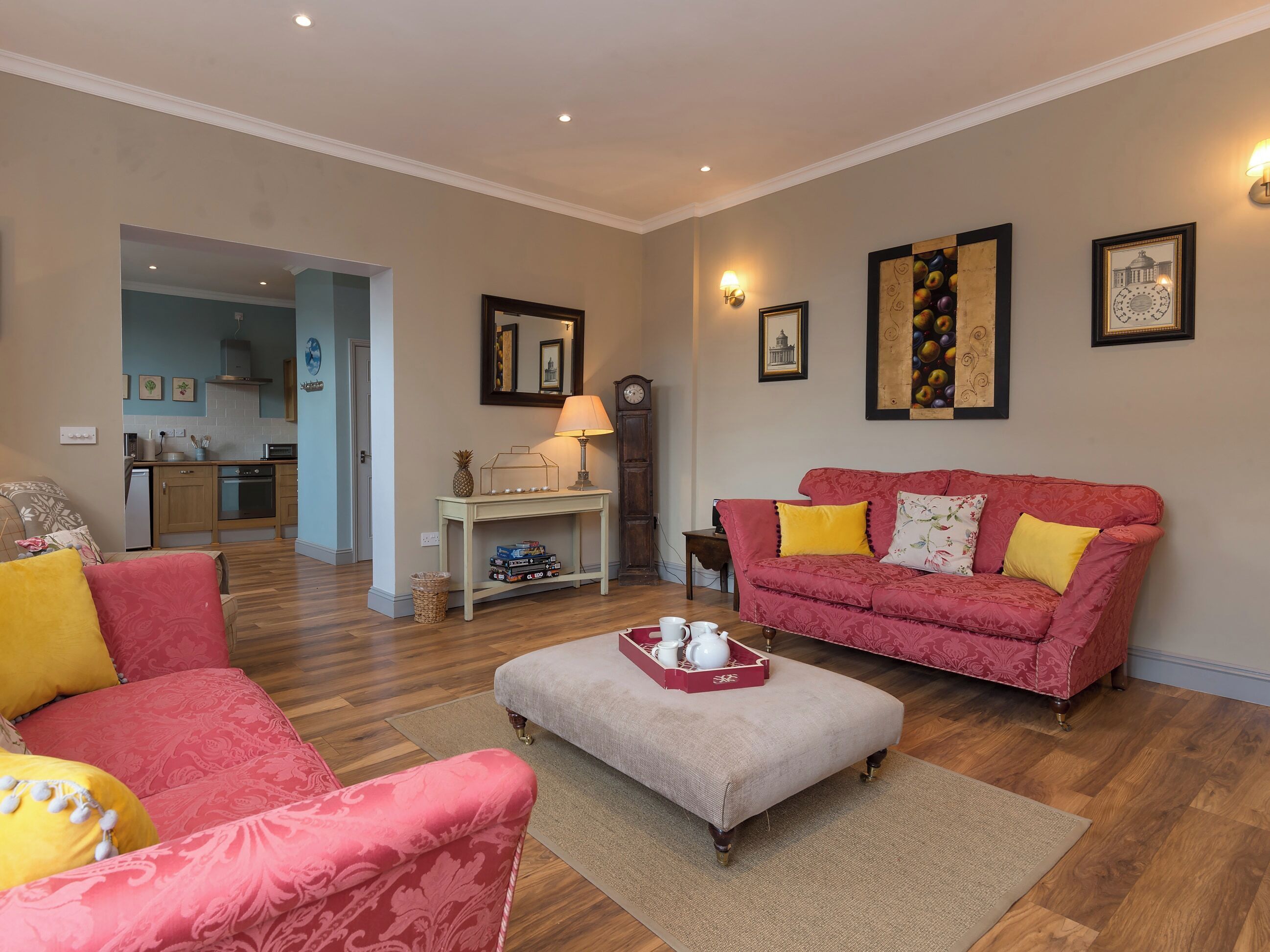 A living room with a red sofa, a grey ottoman, and a coffee table with a tray. There is a kitchen in the background.