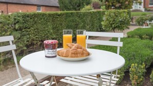 Una mesa blanca con un plato de cruasanes y dos vasos de jugo de naranja, un tarro de mermelada de fresa, y una silla blanca.