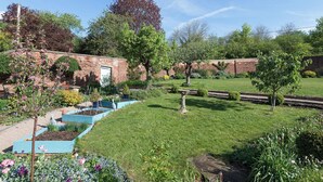 Un jardín con muro de ladrillo, un cobertizo pequeño y diversas plantas y flores.