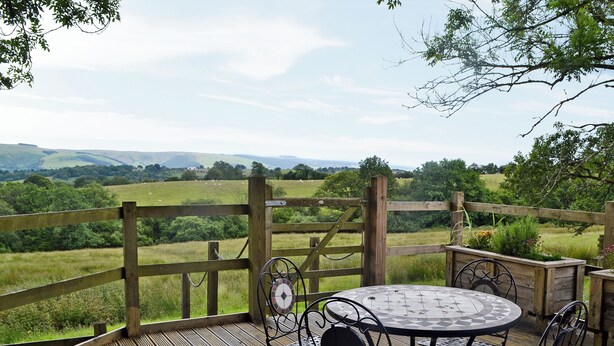 A wooden deck with a round table and chairs, overlooking a grassy landscape with hills. - 1 bedroom accommodation in Garth, near Builth Wells (Garth, near Builth Wells)