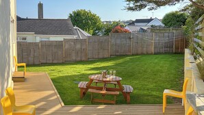 Eine Holzterrasse mit Picknicktisch und gelben Stühlen, umgeben von einem eingezäunten Rasen.