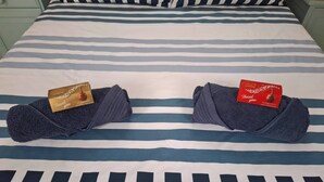 A bed with striped bedding, two folded towels with Lindor chocolate boxes, and a nightstand with a lamp. - Petite Cottage Plett Close to Town (Plettenberg Bay)