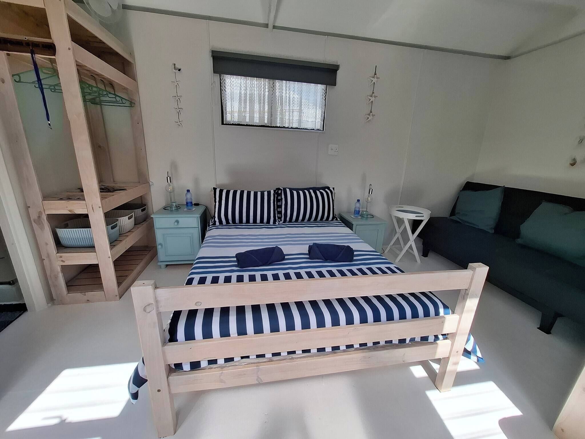 Una habitación con una cama, una mesita de noche, una pequeña mesa, un sofá y una estantería de madera con cestas.