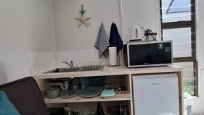 A compact kitchen with a microwave, a small refrigerator, and a sink. There is a window with blinds, a star decoration, and a striped towel hanging on the wall. - Petite Cottage Plett Close to Town (Plettenberg Bay)