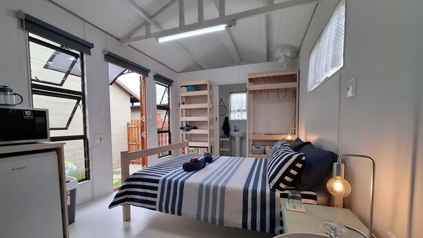 A bedroom with a bed, bedside table, lamp, and a view of the outside. - Petite Cottage Plett Close to Town (Plettenberg Bay)